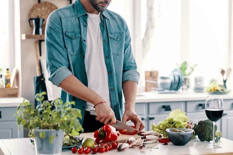 Healthy eating. Close up of young man in casual wear cutting vegetables while standing in the kitchen at home